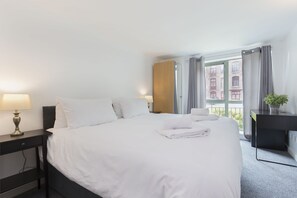 A hotel room with a neatly made bed, a bedside table with a lamp, a desk with a plant, and a window with curtains.