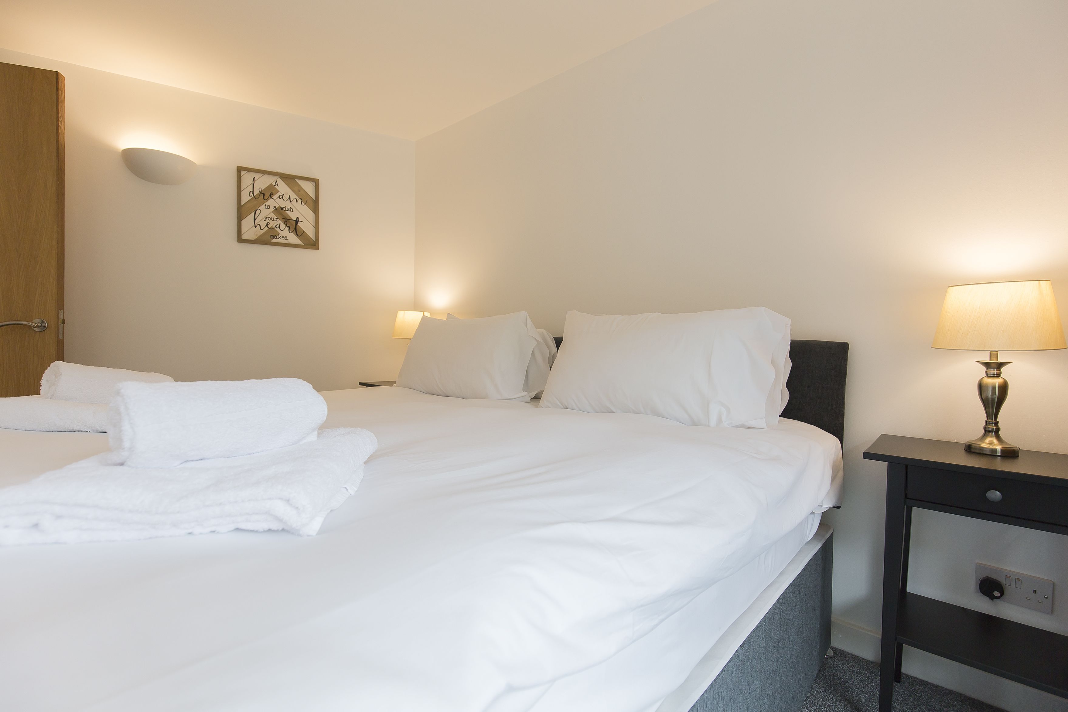 A hotel room with two beds, white linens, a bedside table with a lamp, and a wall-mounted light fixture.