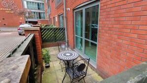 A courtyard with a brick building, a glass door, a small table with chairs, and a parked car. - Raleigh Square Apartments (Nottingham)