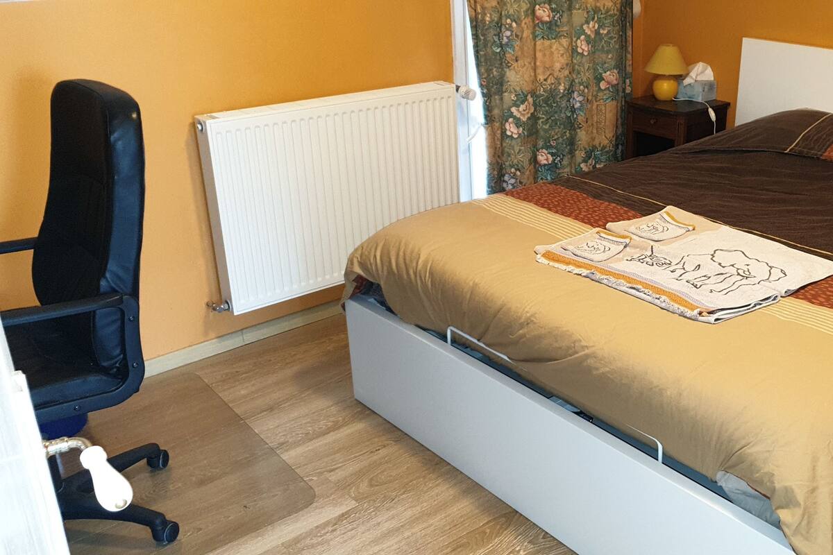 A neatly made bed with a brown and beige bedspread, a small decorative pillow, and a folded blanket with a design. There is a black office chair with wheels, a white radiator, and a nightstand with a lamp and a tissue box.