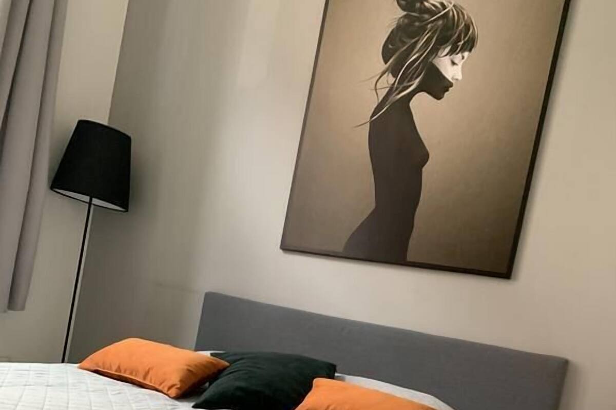 Un lit bien fait, avec une couette blanche matelassée et deux oreillers, un orange et un vert, dans une chambre moderne dotée d’une tête de lit grise et d’un lampadaire.