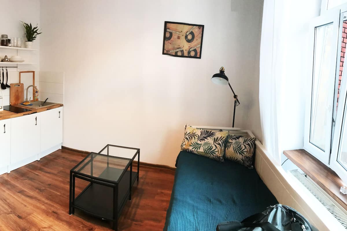 Un espace de vie compact comprenant un coin cuisine, une table basse en verre, un canapé avec des coussins à motifs et une vue sur la ville par la fenêtre.