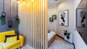 A modern bedroom with a wooden slat partition, a yellow sofa, a bed with a white headboard, and wall-mounted shelves with framed artwork. - ApartMood (Katowice)