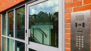 A building entrance with a glass door, a visible staircase, and a metal intercom system. - Portland Square (Nottingham)