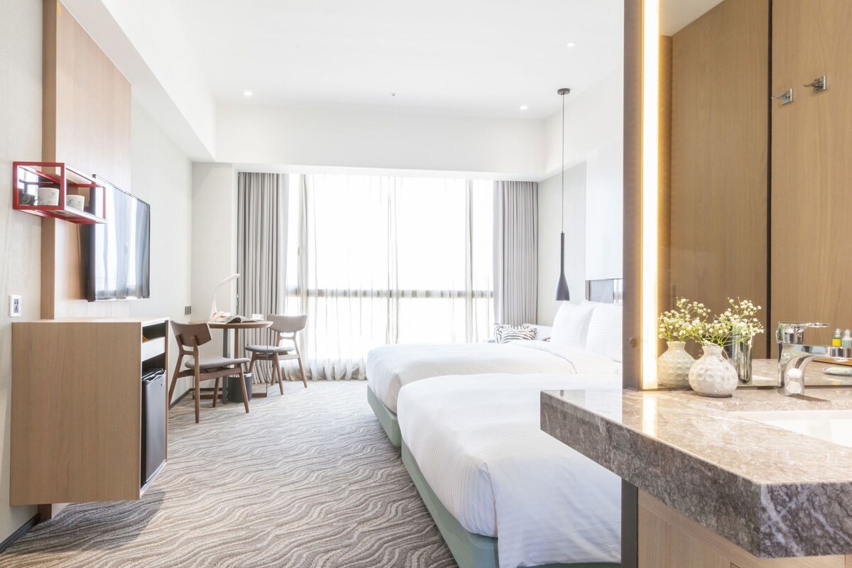 Habitación de hotel con cama, escritorio, silla y un ventanal con cortinas.