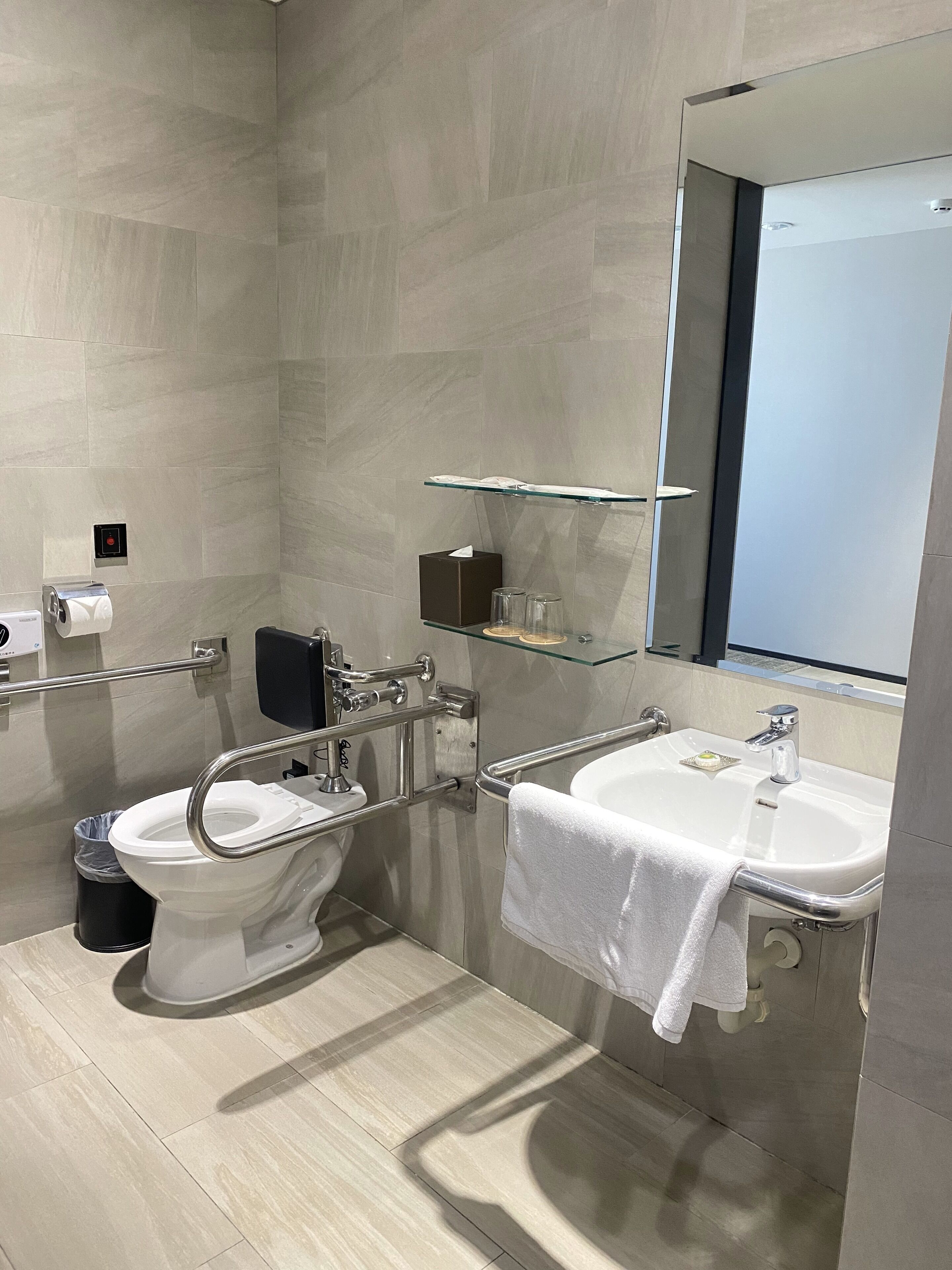 Un baño moderno con inodoro, bidé y lavamanos, equipado con barras de apoyo y un estante de vidrio sobre el lavamanos.