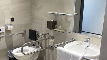 Un baño moderno con inodoro, bidé y lavamanos, equipado con barras de apoyo y un estante de vidrio sobre el lavamanos.