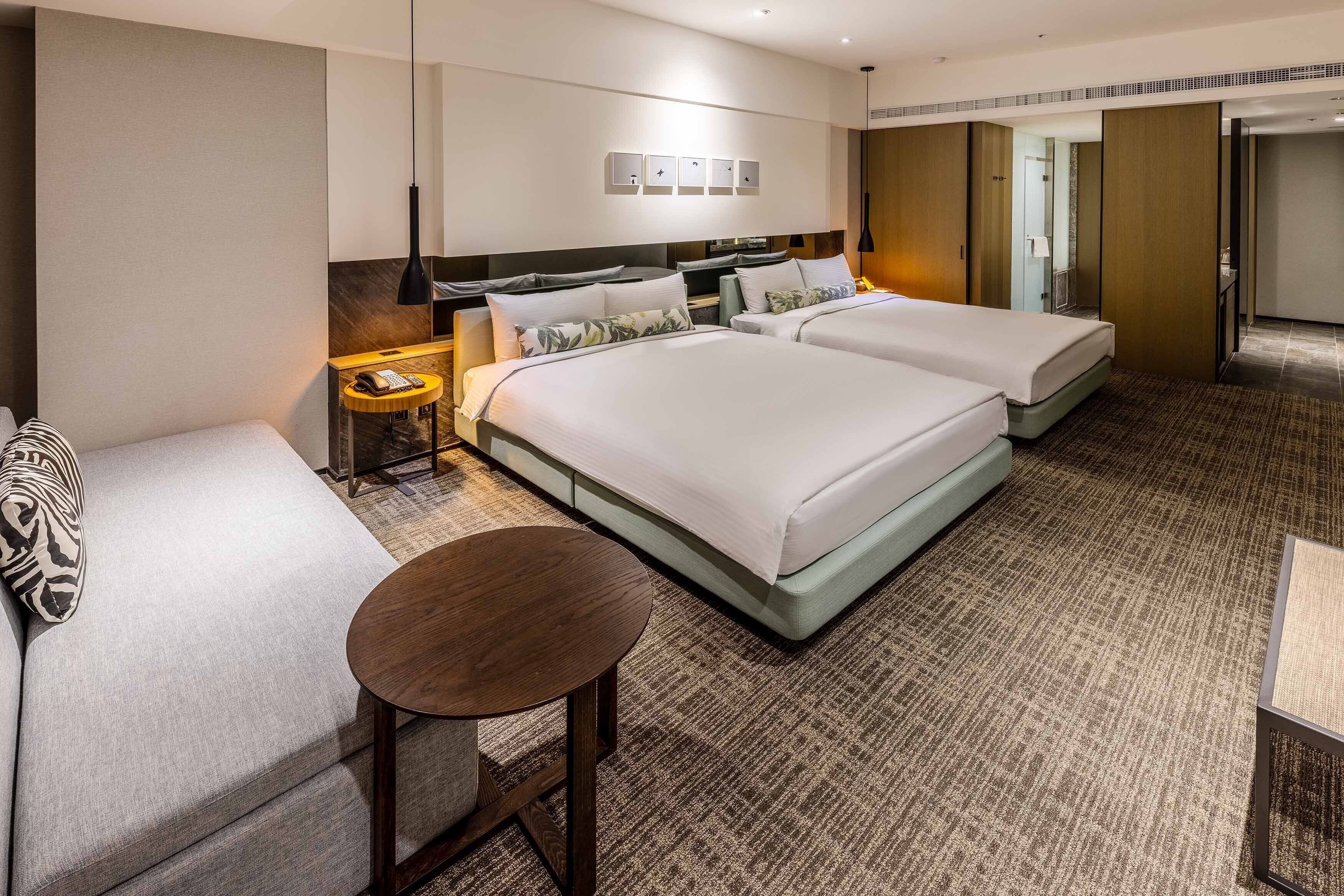 A hotel room with two beds, a sofa, a small round table, and a nightstand with a lamp.