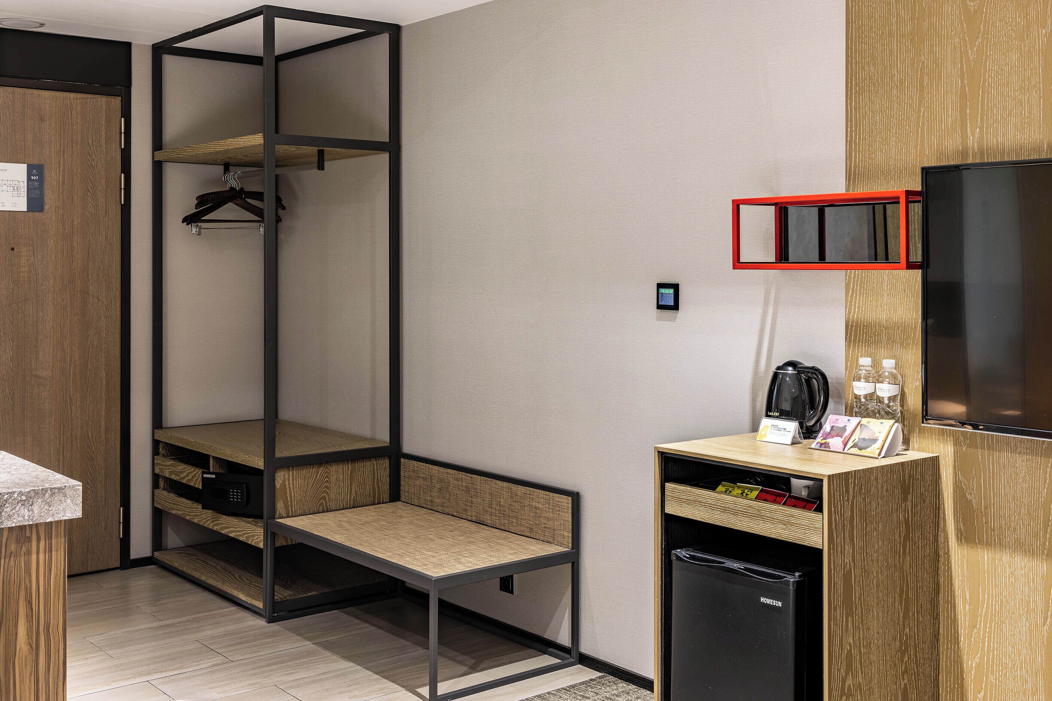 Ein modernes Hotelzimmer mit einem Holzschrank, einer eingebauten Sitzbank, einer kleinen Küchenzeile inklusive Kaffeemaschine und einem an der Wand montierten Fernseher.