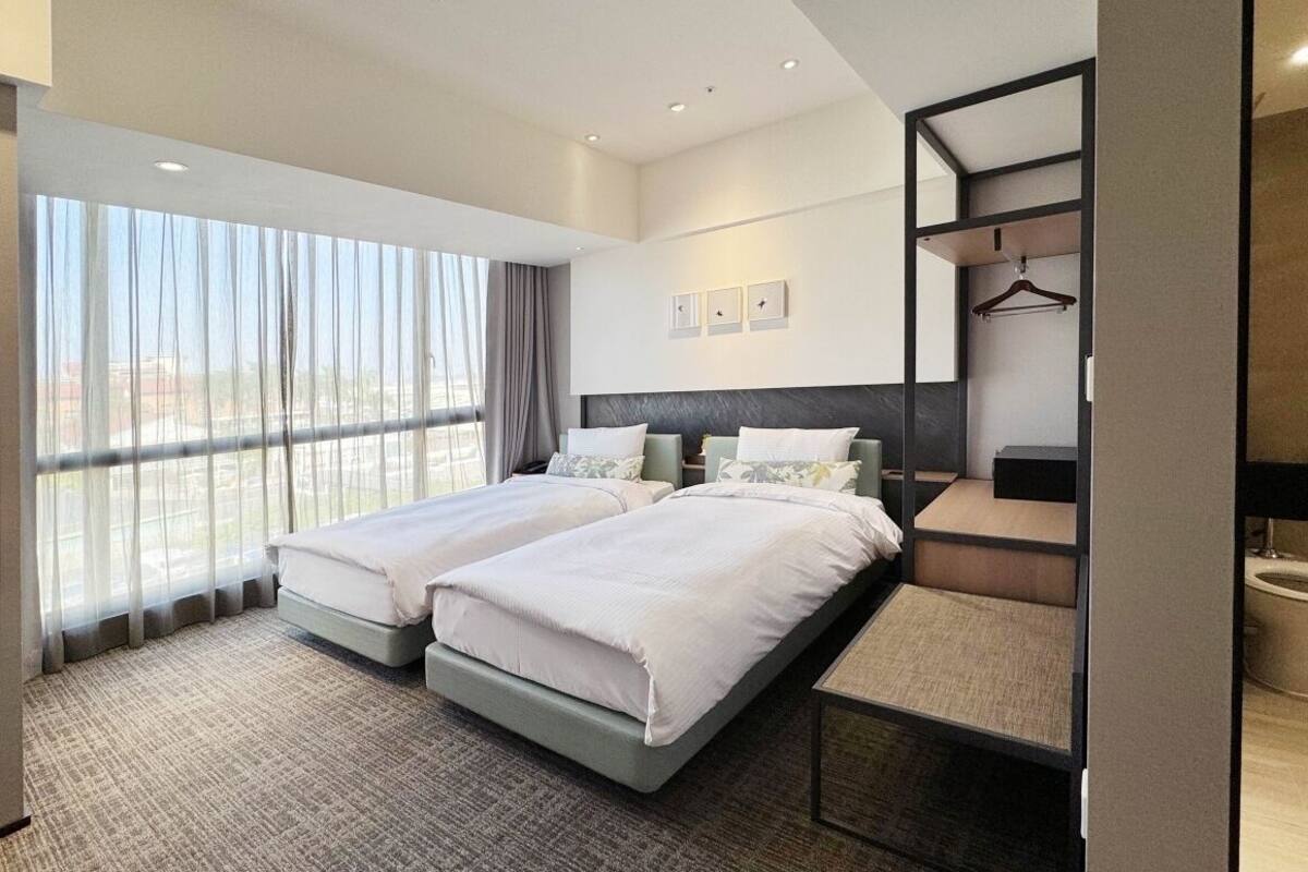 A hotel room with two beds, a large window with sheer curtains, a mirror, and a small table.