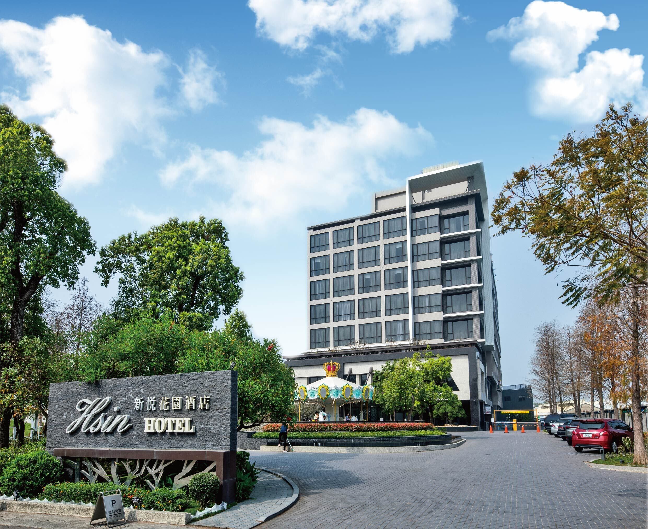 A hotel entrance with a sign, a parking area, and a modern building in the background.