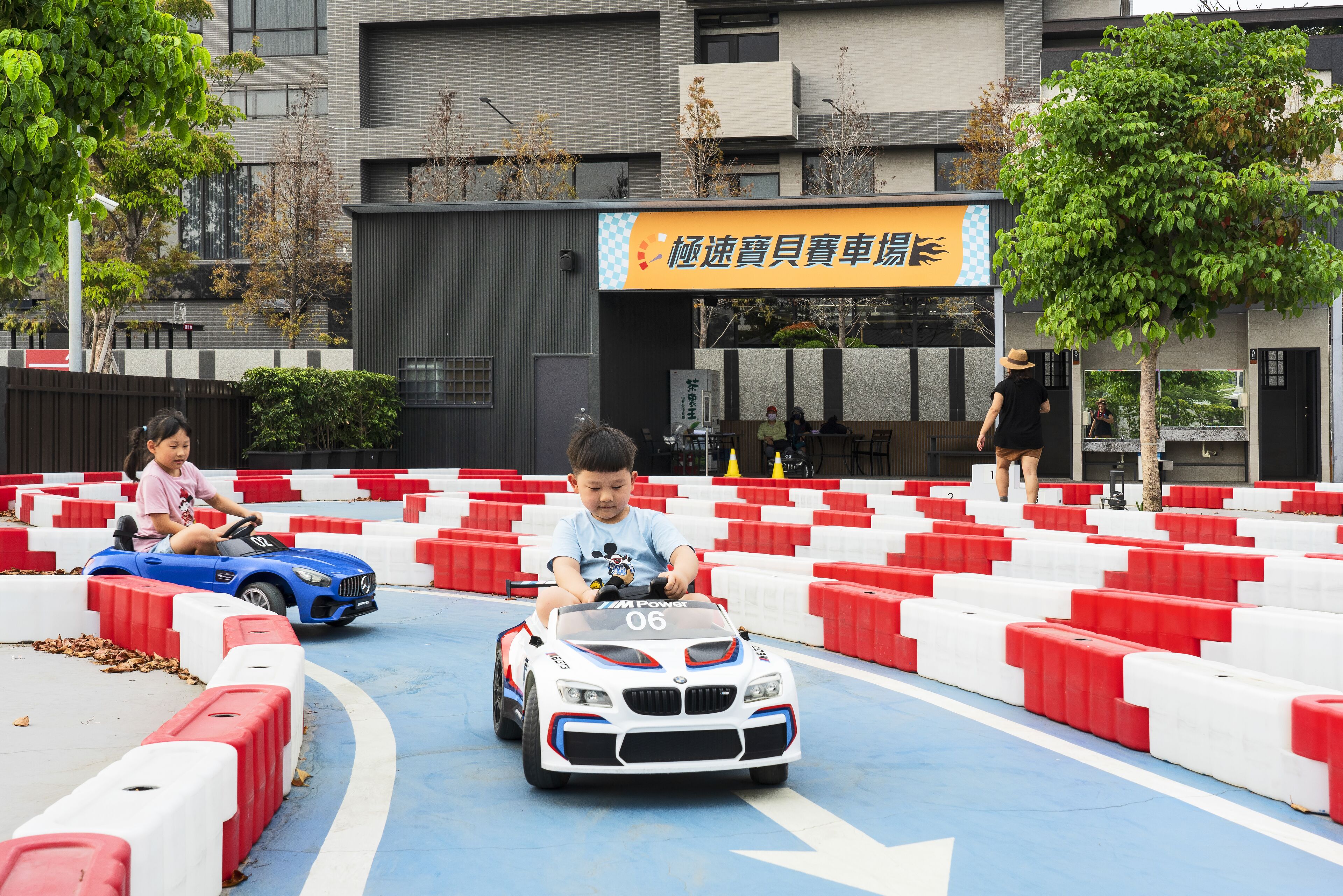 Children driving toy cars on a track with red and white barriers.