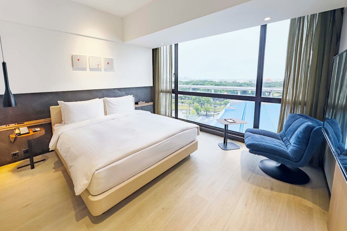 A modern hotel room with a large bed, a desk, a chair, and a view of a bridge and cityscape through large windows.