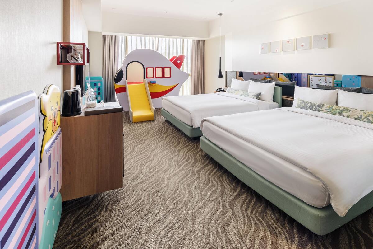 A hotel room with two beds, a desk with a computer, a children's play area, and a large window with curtains.