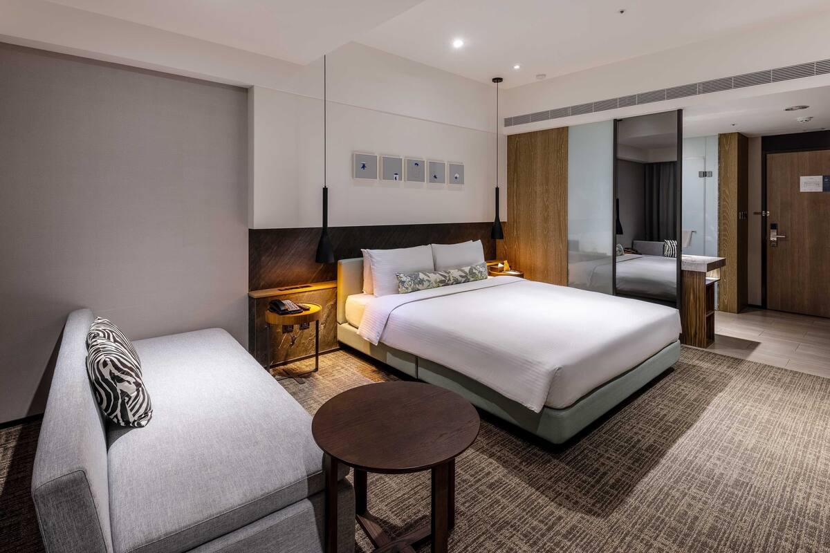 A modern hotel room with a large bed, a sofa, a small round table, and a bathroom visible through a sliding door.