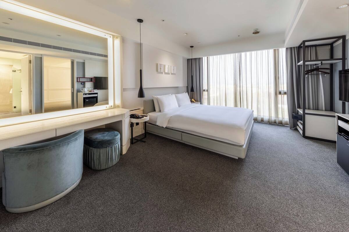 A modern hotel room with a large bed, a desk, a chair, a mirror, a wardrobe, and a TV.