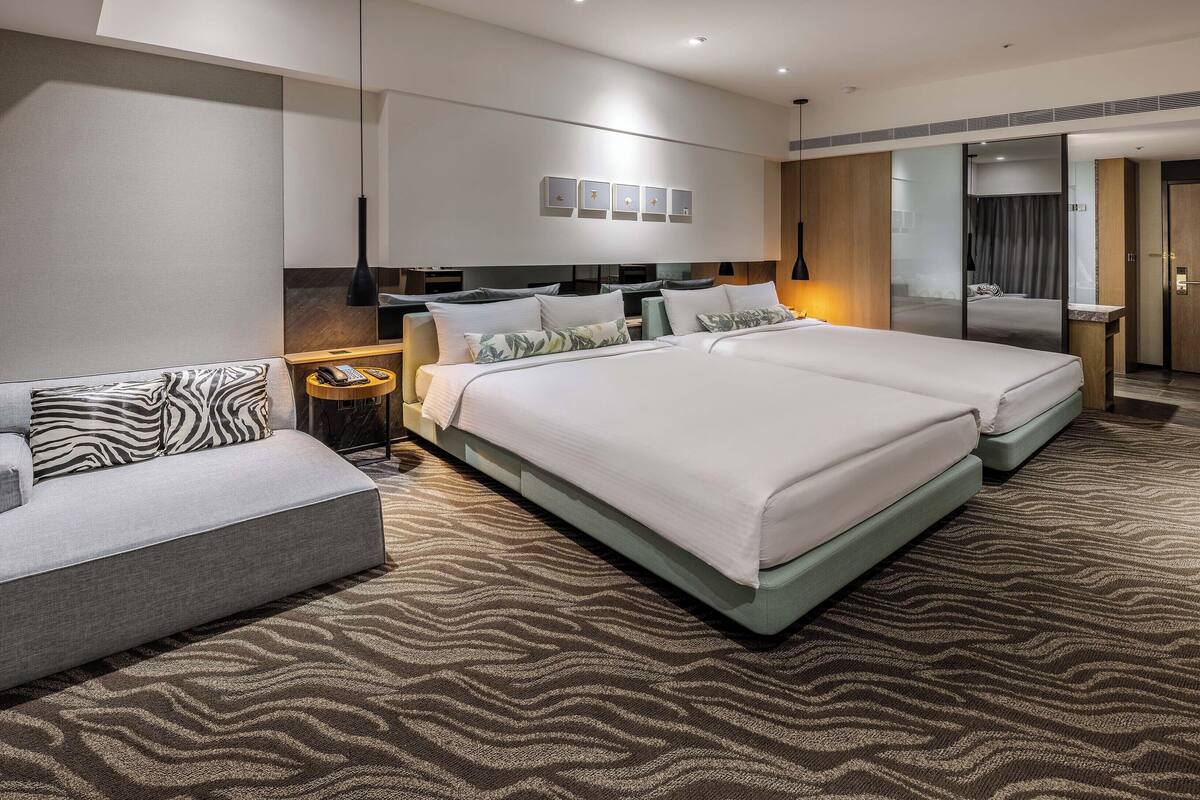 A modern hotel room with a large bed, a sofa, a bedside table, a mirror, and a wall-mounted light fixture.