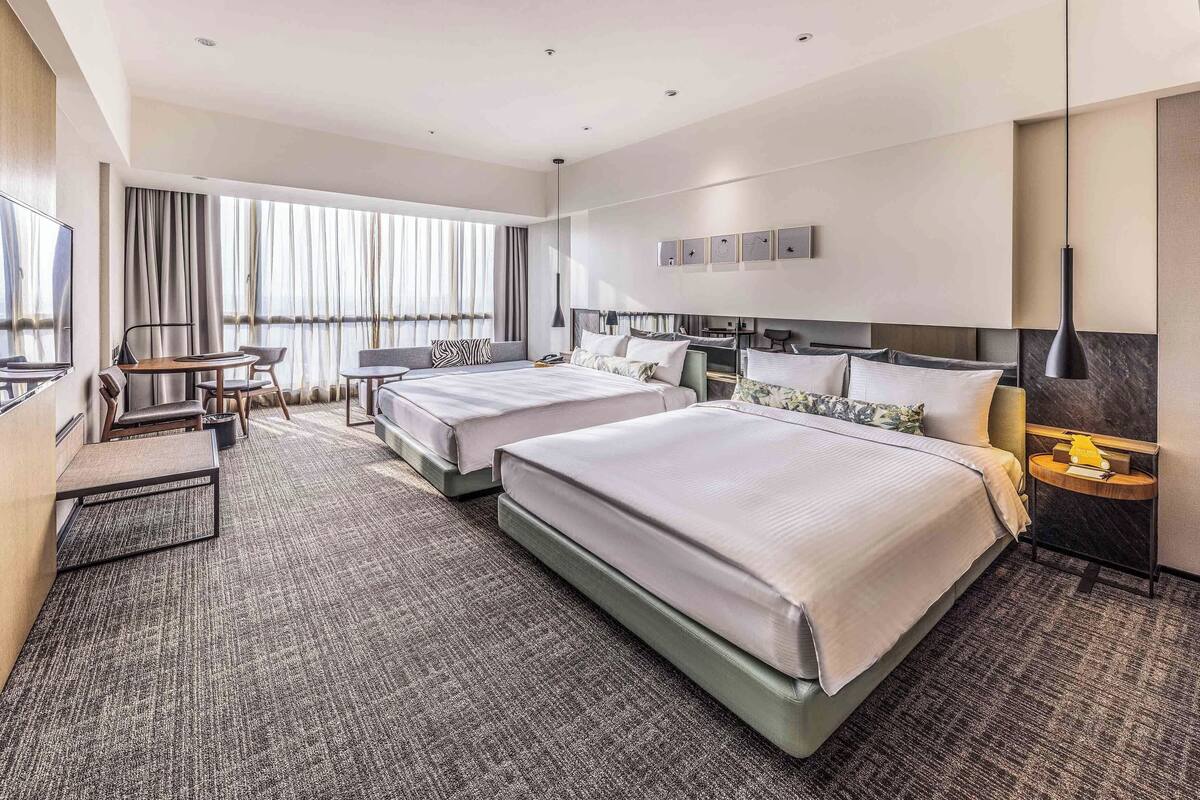 A hotel room with two beds, a desk, a chair, a TV, and a large window with curtains.