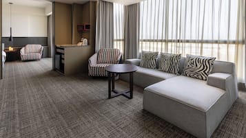 A hotel room with a grey sofa, patterned armchair, and a small round table. There is a bar area with a mirror and a shelf with items. The room has large windows with curtains.