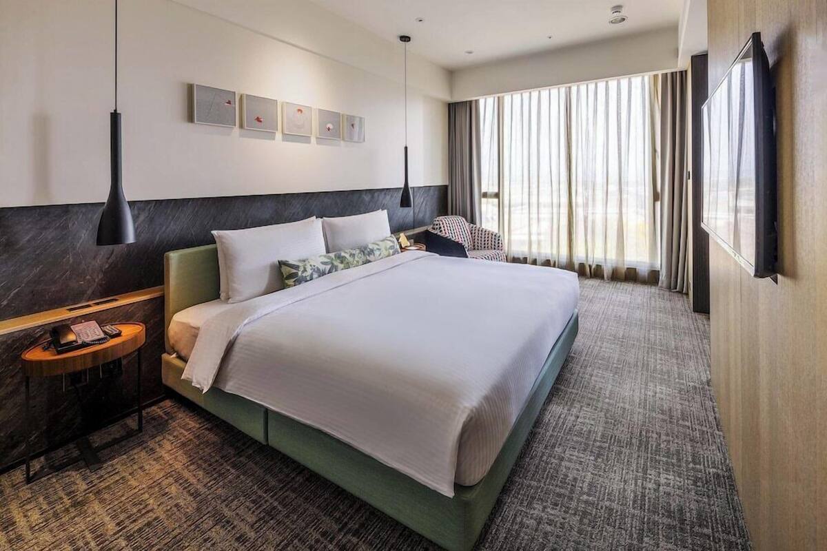 A hotel room with a large bed, a bedside table with a telephone, a wall-mounted TV, and a large window with curtains.