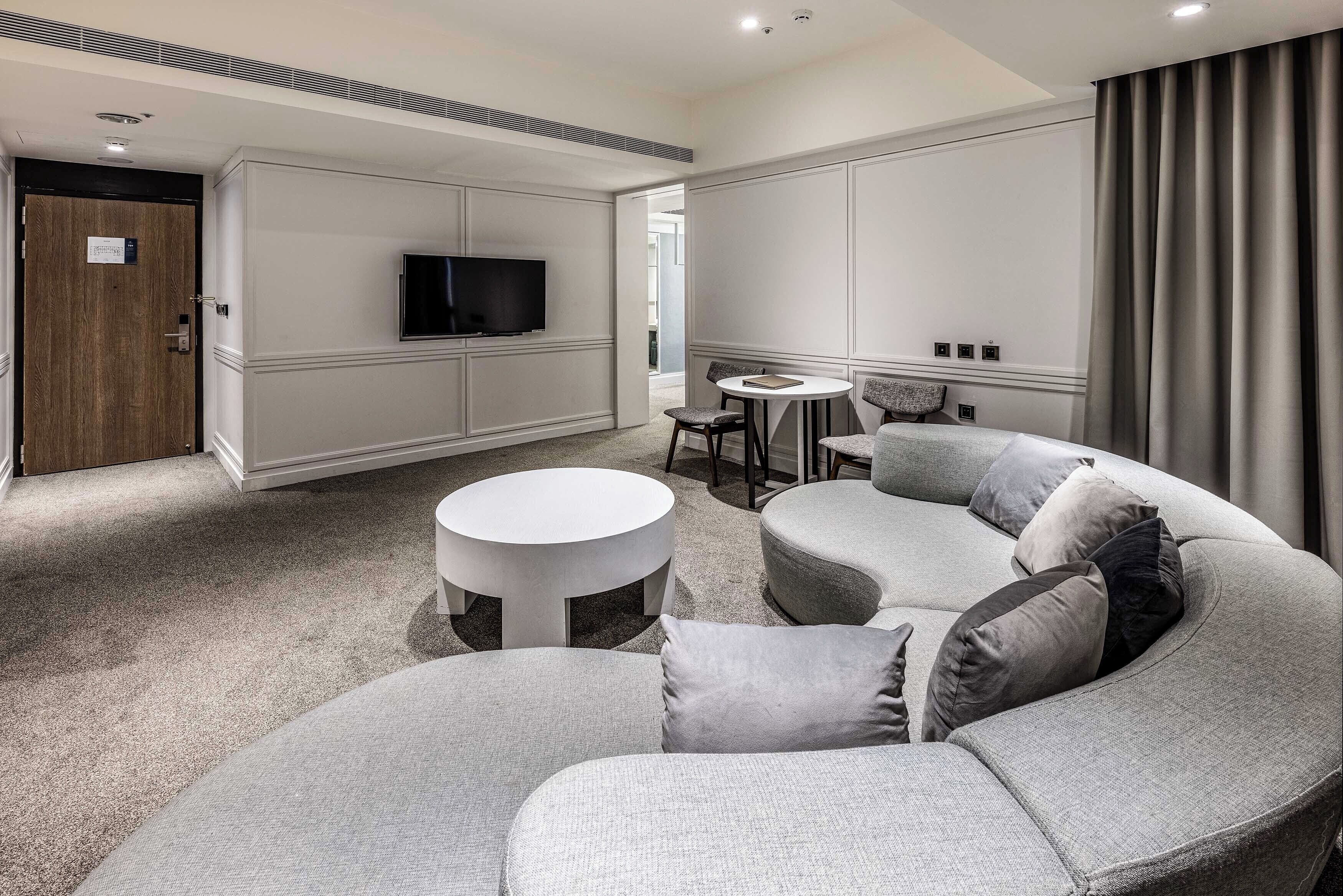 A modern hotel room with a curved grey sofa, a round white coffee table, a flat-screen TV mounted on the wall, and a small round table with chairs.
