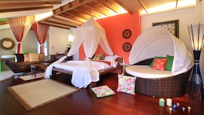 Un dormitorio con una cama bajo un dosel, una silla de mimbre con cojines, una mesita y un espejo decorativo en la pared.