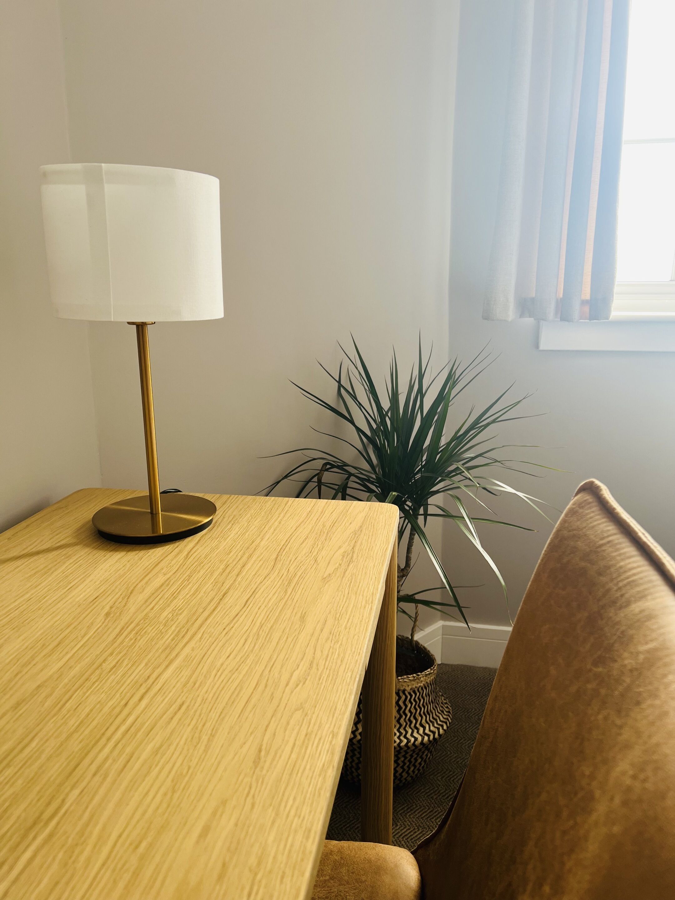 A wooden table with a gold lamp, a potted plant, and a brown couch in a room with a window and curtains.