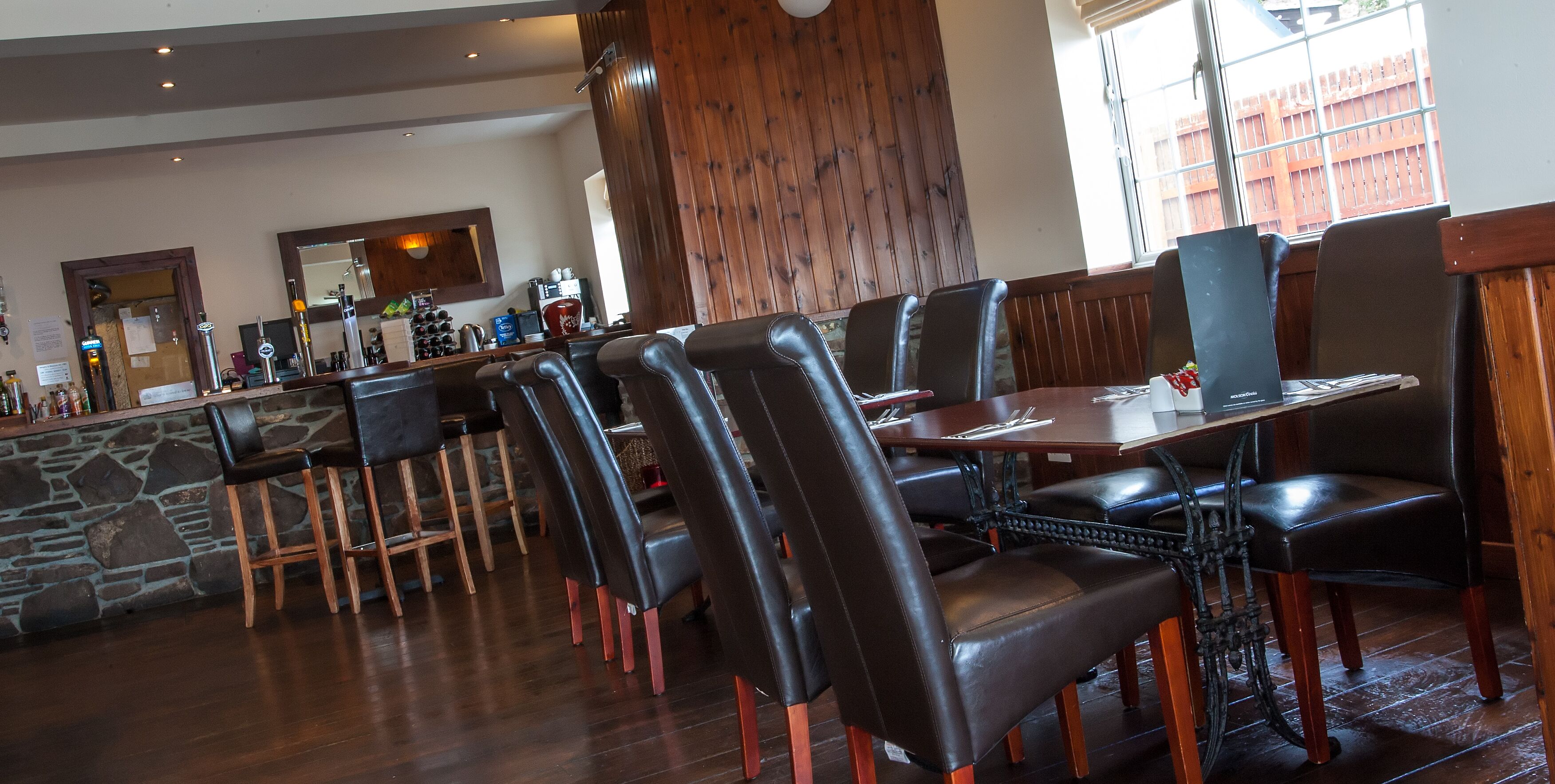 A restaurant with wooden flooring, leather chairs, a stone wall, and a bar area with various bottles and glasses.