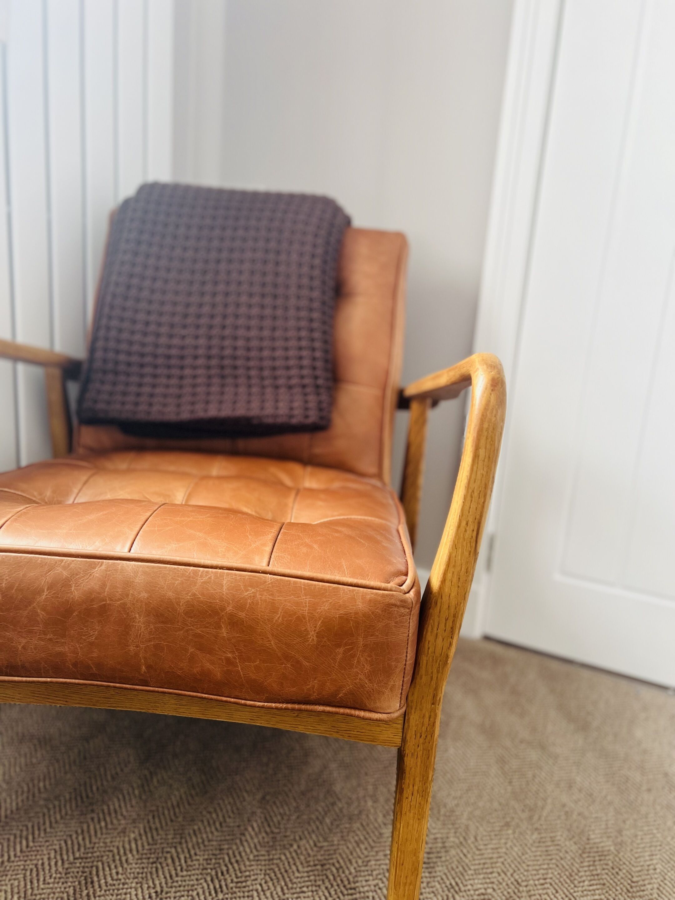 A brown leather chair with wooden armrests and a cushion, positioned against a white wall and carpeted floor.