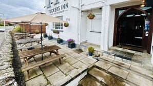 Un bâtiment blanc avec une enseigne indiquant « Tuckers Inn, hôtel, bar, restaurant ». On y trouve des tables et des bancs en bois à l’extérieur, des plantes en pot et un chemin de pierre menant à l’entrée.