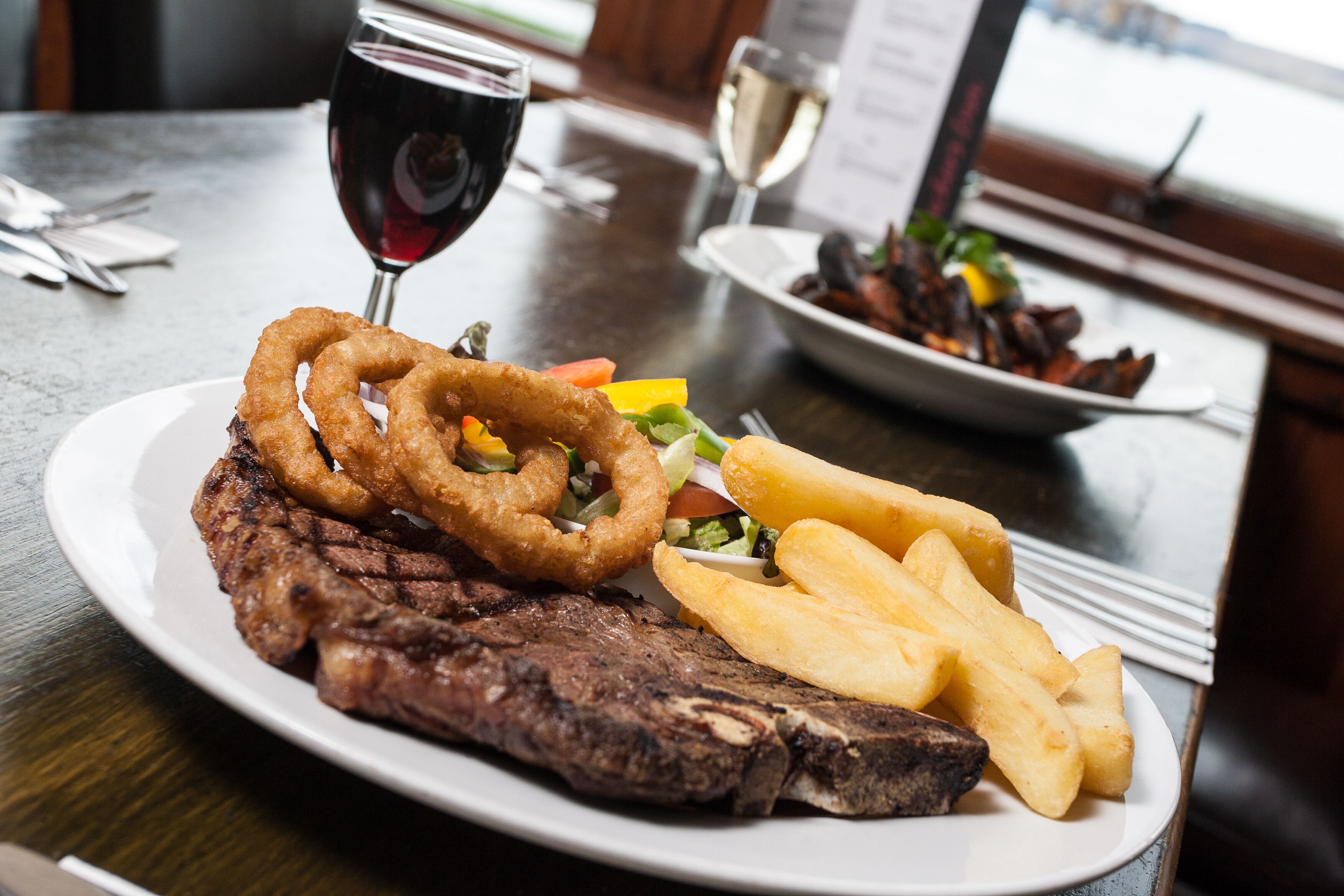 Un plato de carne con aros de cebolla, papas fritas y ensalada, acompañado de una copa de vino tinto y una copa de vino blanco sobre una mesa de madera.