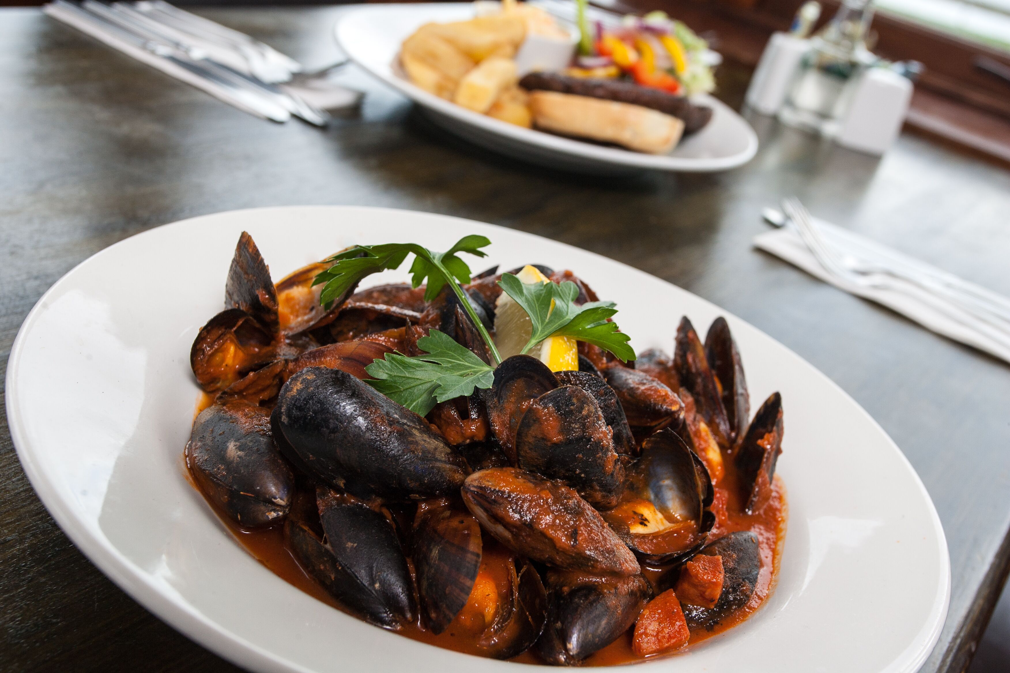A plate of mussels in a red sauce, garnished with parsley, on a dark table with a napkin and utensils in the background.