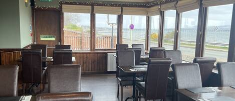 A restaurant with wooden flooring, leather chairs, and tables. There are large windows with a view of a bridge and a body of water.