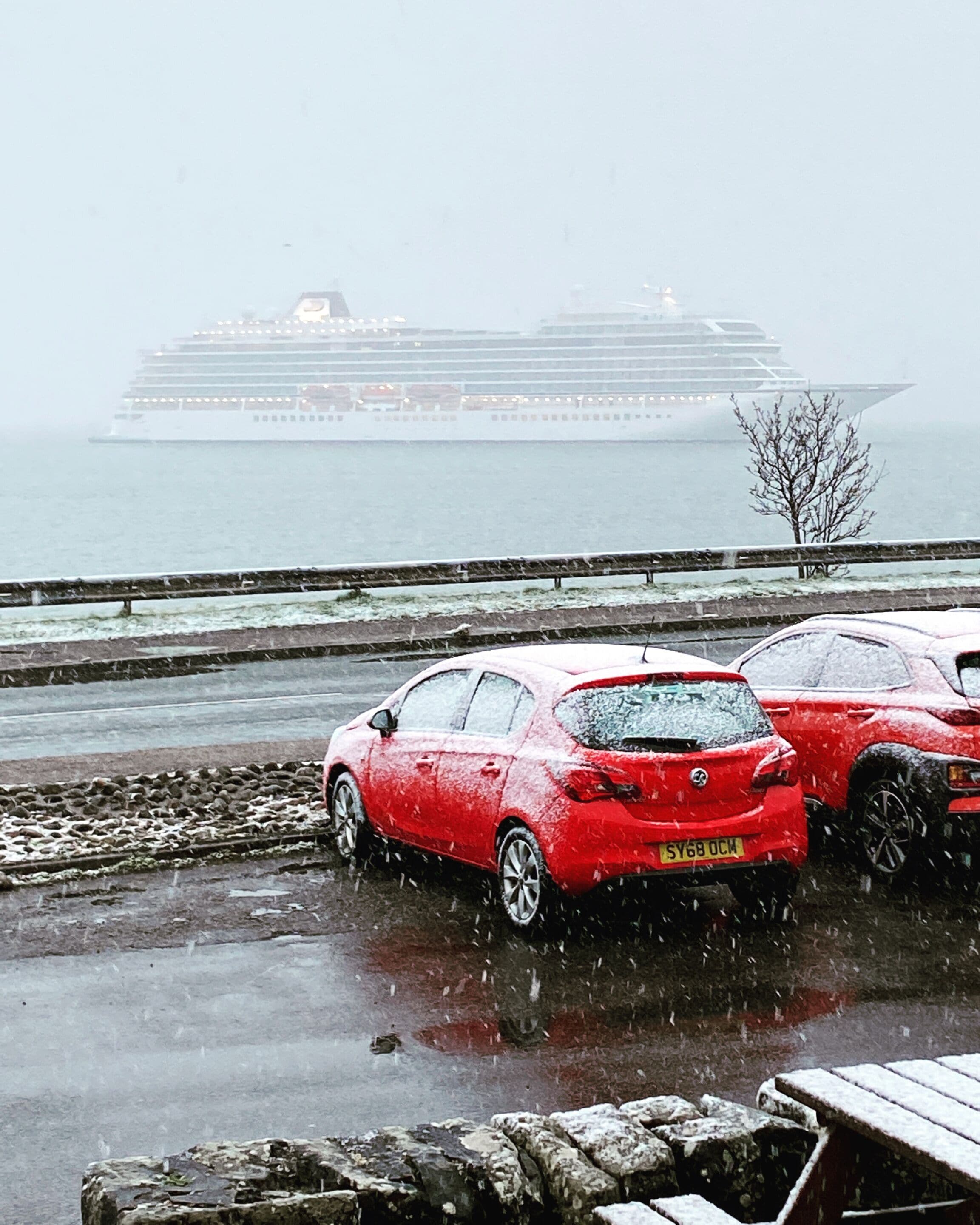 A red car parked on a wet surface with a cruise ship in the background.