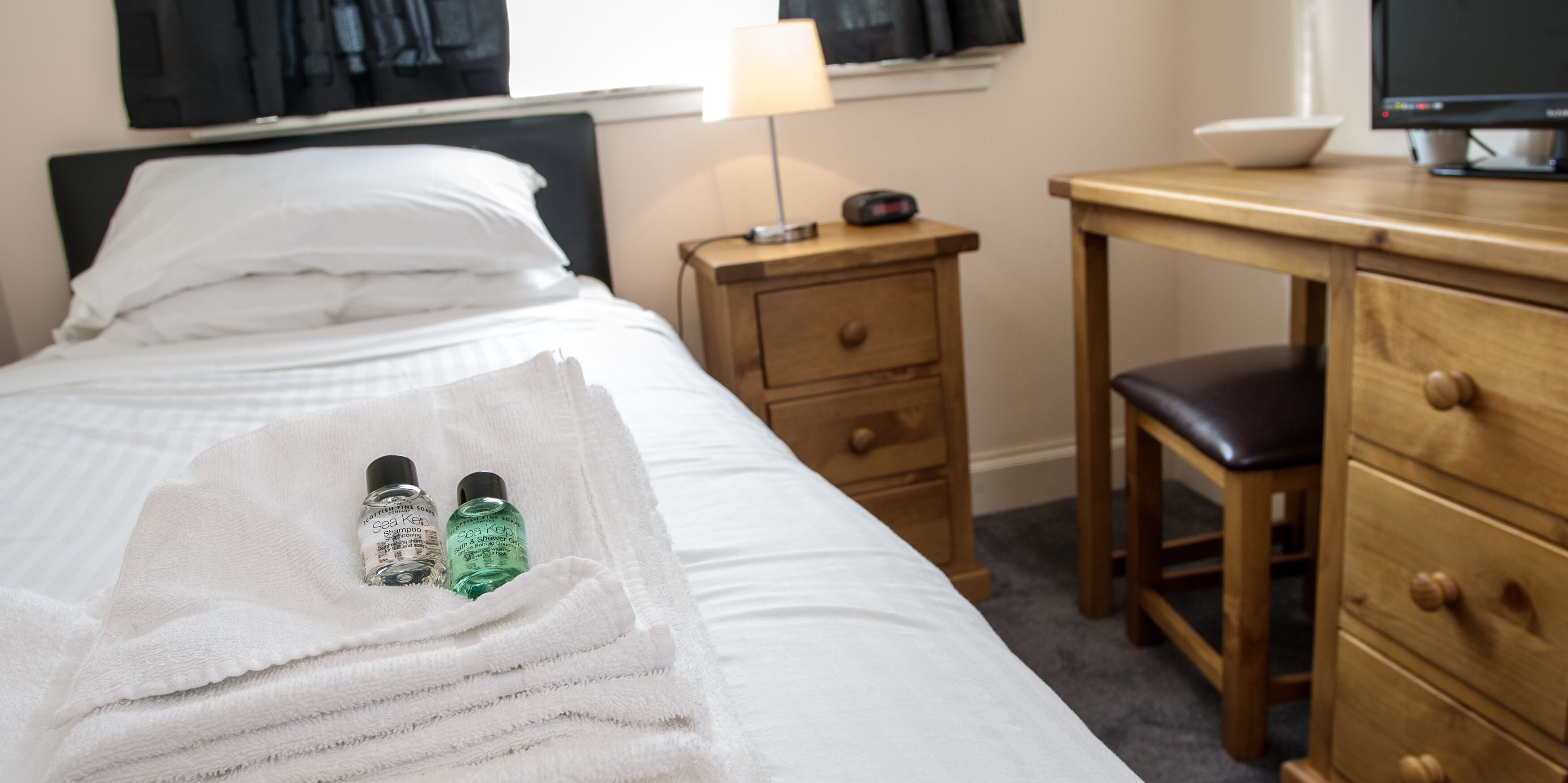 A neatly made bed with a white towel and two bottles of shampoo. There is a wooden nightstand with a lamp and a clock beside the bed. A wooden desk with a chair is also visible.