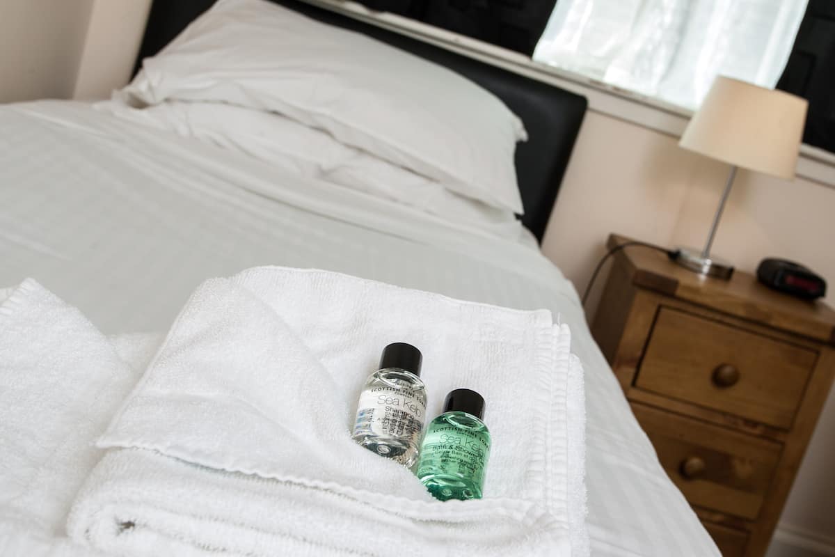 A neatly made bed with white linens, a wooden nightstand with a lamp, and two bottles of Sea Kelp shampoo on the bed.