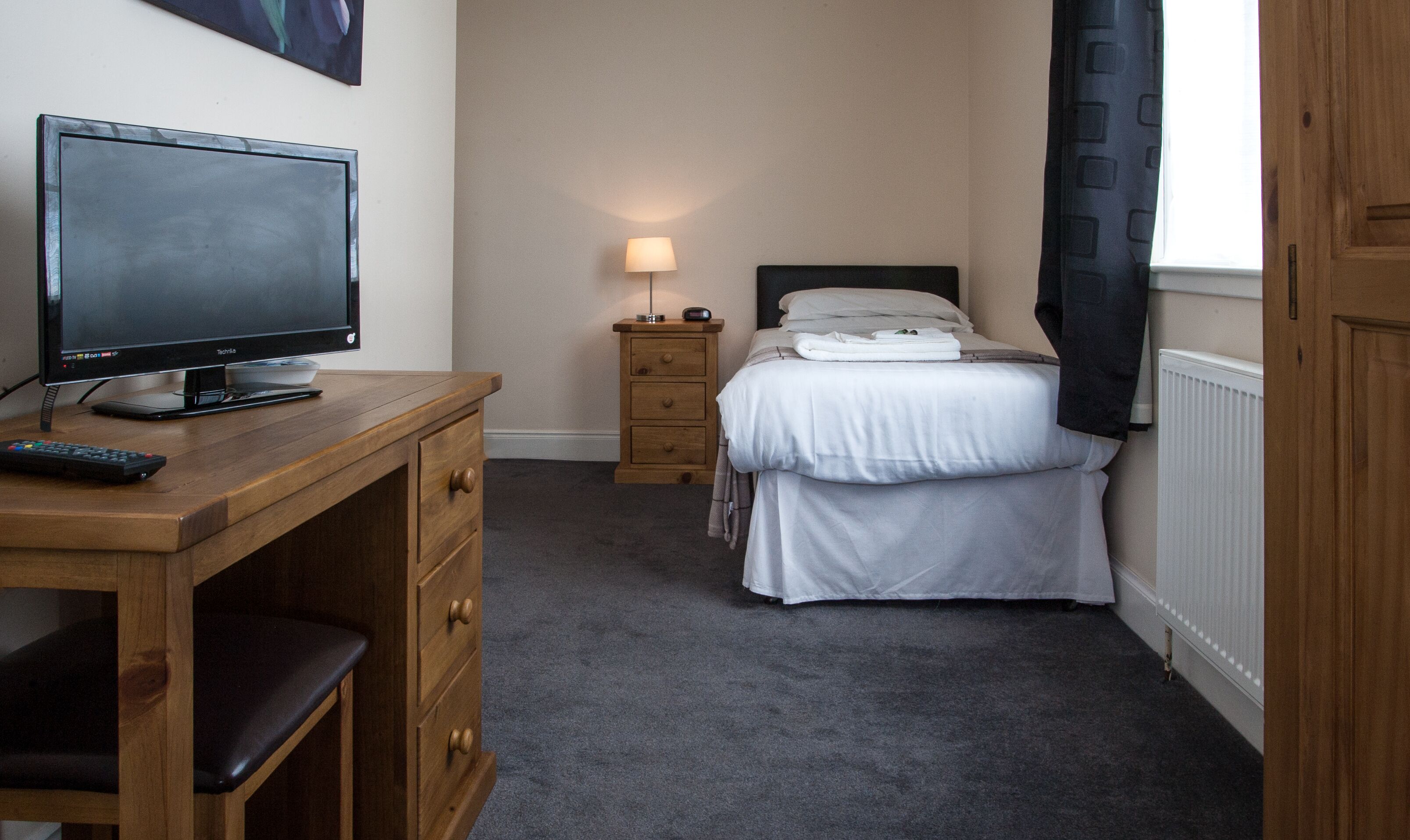 A hotel room with a bed, a bedside table with a lamp, a TV on a wooden desk, and a window with curtains.