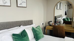 Un lit bien fait, avec du linge de lit blanc et des coussins verts, un bureau en bois avec un miroir, et deux illustrations botaniques encadrées accrochées au mur.