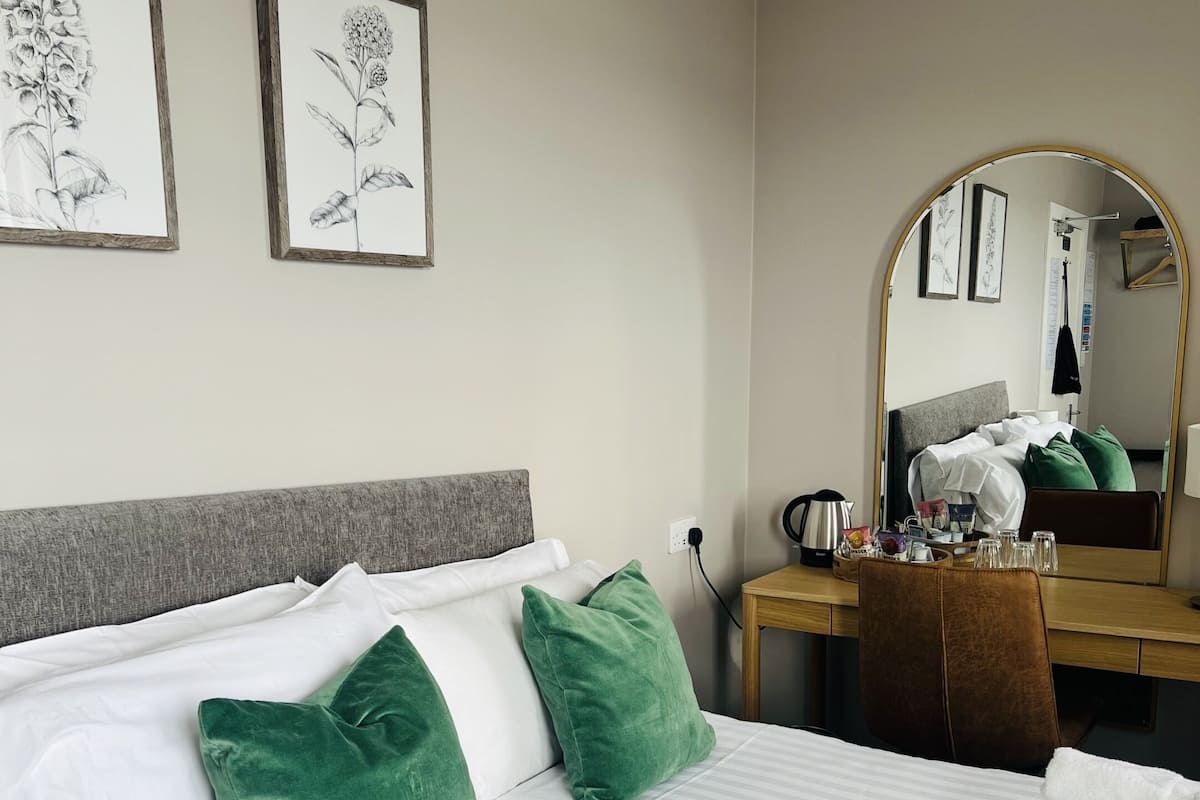 A neatly made bed with white linens and green pillows, a wooden desk with a mirror, and two framed botanical illustrations on the wall.