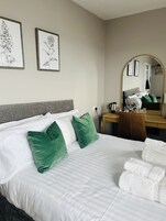 A neatly made bed with white linens and green pillows, a wooden desk with a mirror, and two framed botanical illustrations on the wall.