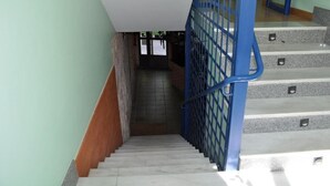 Un escalier avec une rampe bleue et une rampe en bois sur le côté gauche, menant à un espace intérieur doté d’un sol carrelé et d’une porte en bas.