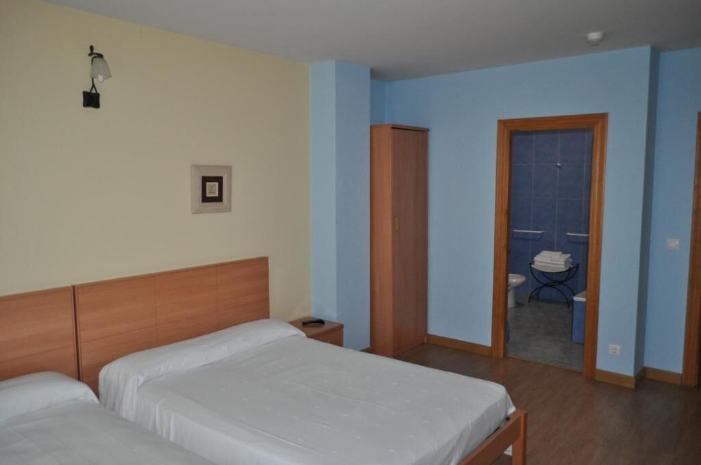 Habitación de hotel con dos camas, un armario de madera, una mesita de noche pequeña, una lámpara de pared y un baño visible a través de una puerta abierta.