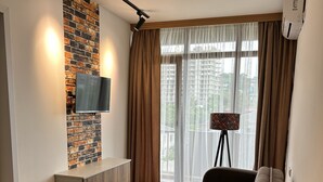 Un salon moderne avec une table à manger en bois, un canapé, un téléviseur fixé à un mur en briques et une grande fenêtre avec des rideaux.