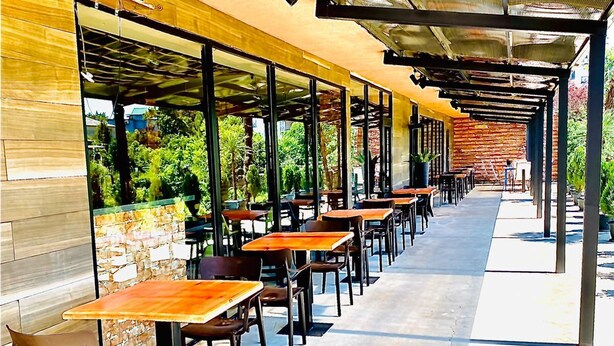 Un café en plein air avec des tables et des chaises en bois, un mur en briques et un espace de repos couvert avec un toit en métal.