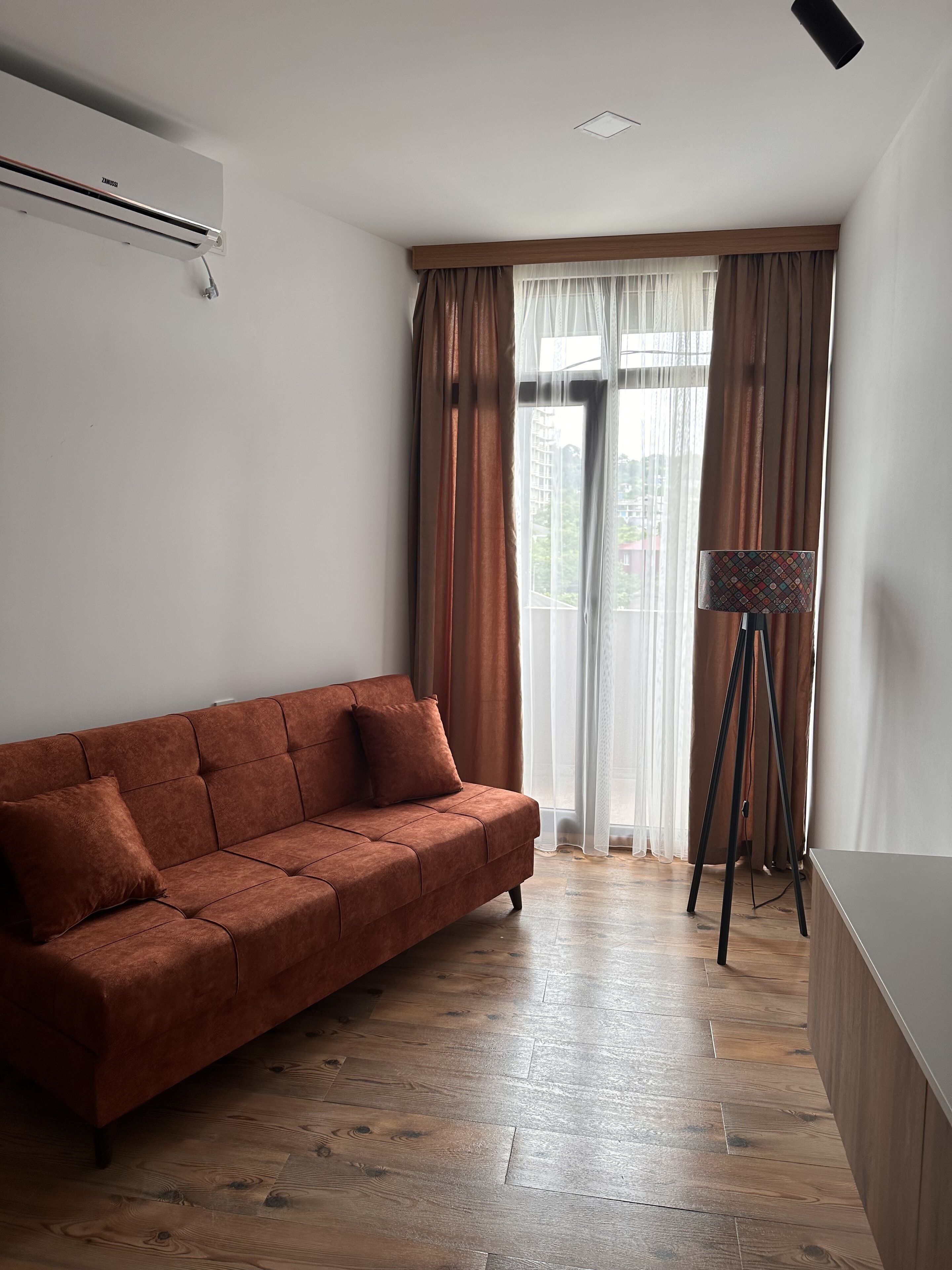 A room with a brown sofa, a standing lamp, and a window with curtains.