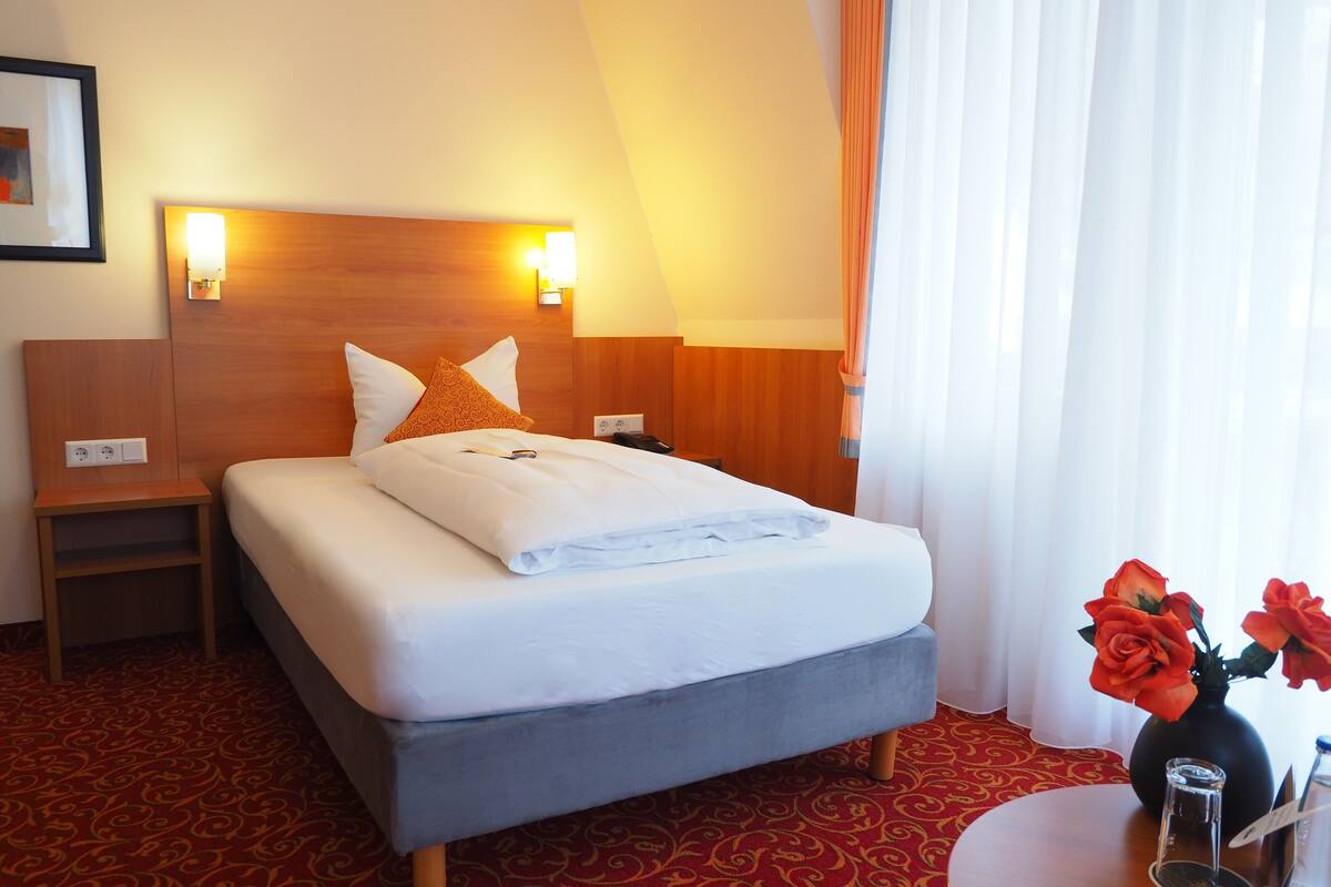Ein Hotelzimmer mit Bett, Nachttischen, einer Lampe, einer Vase mit Blumen und einem Fenster mit Vorhängen.
