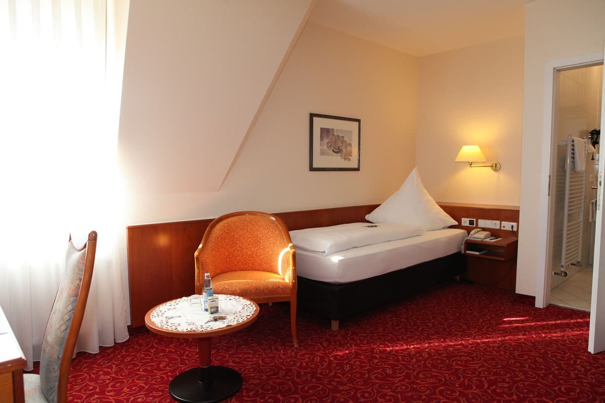 Ein Hotelzimmer mit einem Bett, einem Sessel, einem kleinen runden Tisch, einer Wandlampe und einem gerahmten Bild.