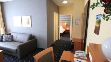 Et hotelværelse med en sofa, et skrivebord med telefon og papirer, en seng i baggrunden og en dør, der fører til et andet rum.