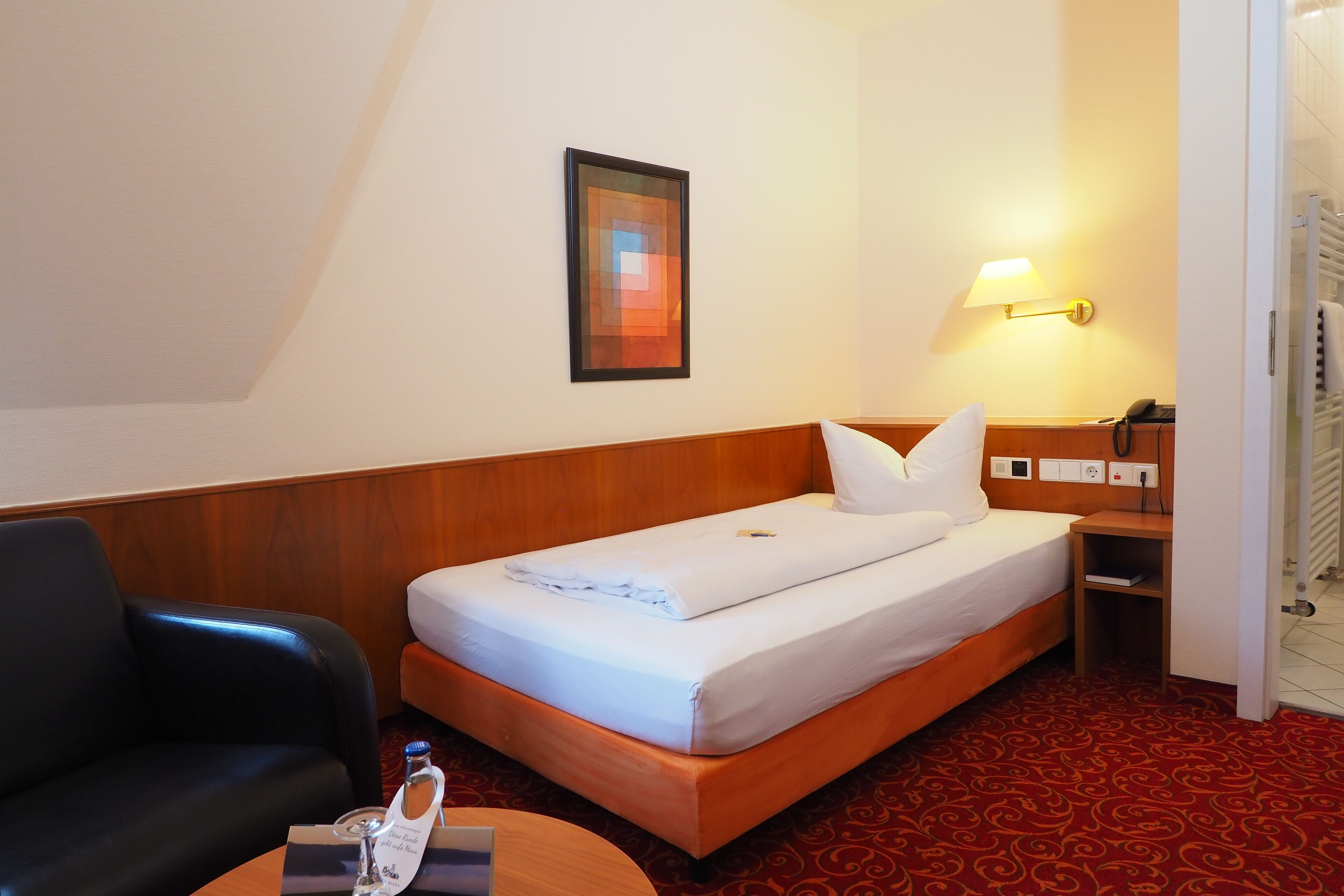 Een hotelkamer met een enkel bed, een houten hoofdeinde, een nachtkastje met lamp, een zwarte leren fauteuil en een vloerkleed met een rood patroon.