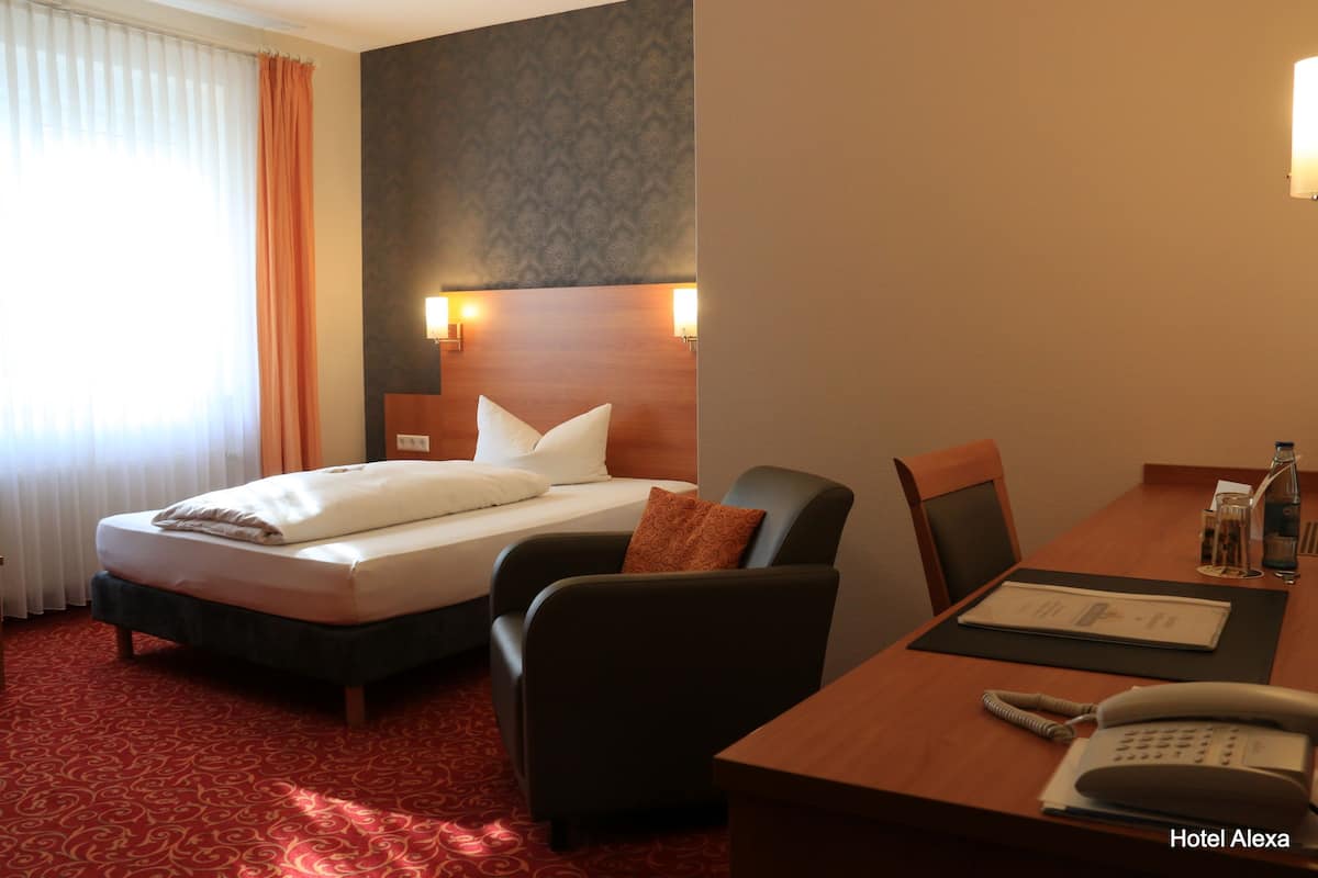 Ein Hotelzimmer mit einem Bett, einem Schreibtisch und einem Stuhl. Auf dem Schreibtisch steht ein Telefon und auf dem Tisch eine Wasserflasche.